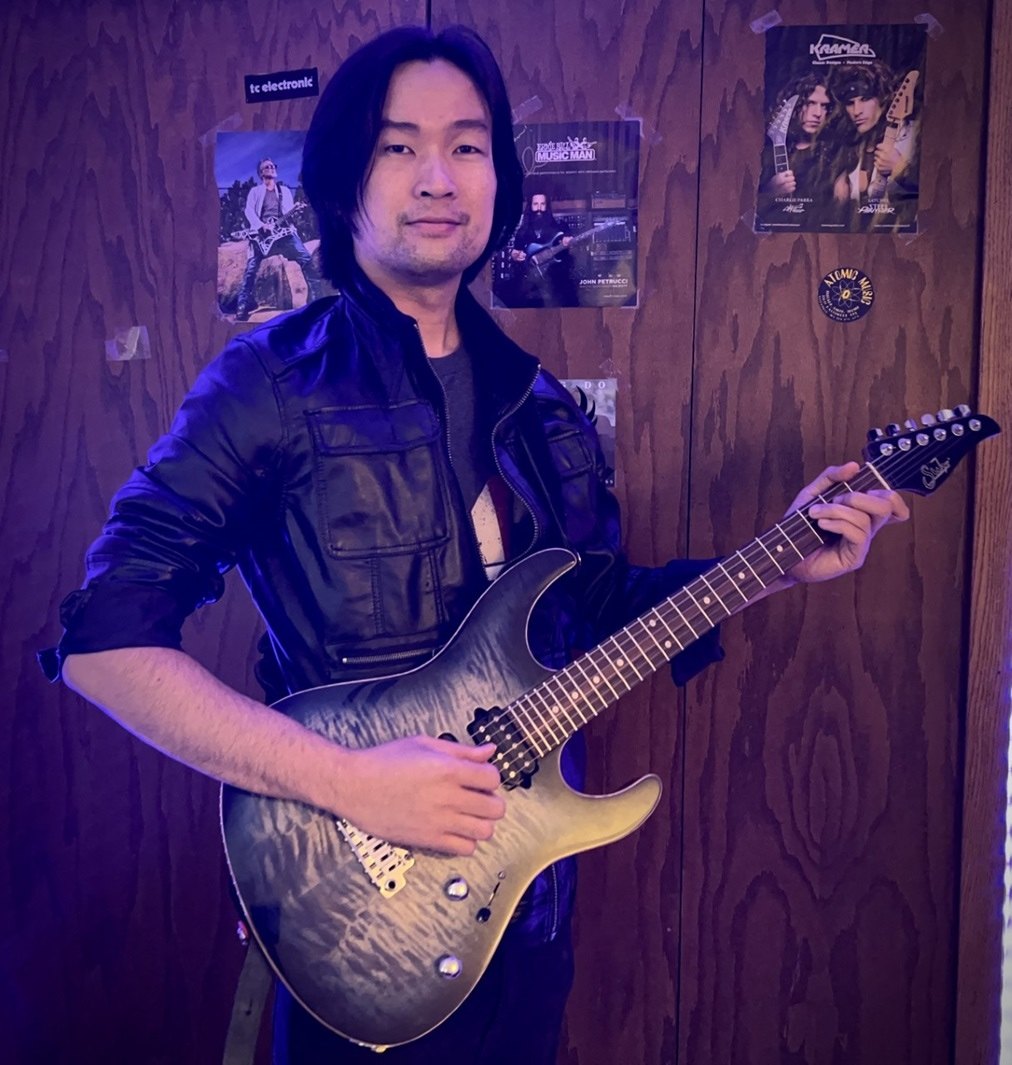 Bang Do holding a teal electric guitar in front of a wall of concert posters