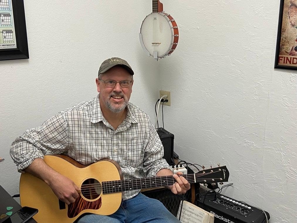 Guitar teacher playing an acoustic in a lesson room with a banjo on the wall behind him