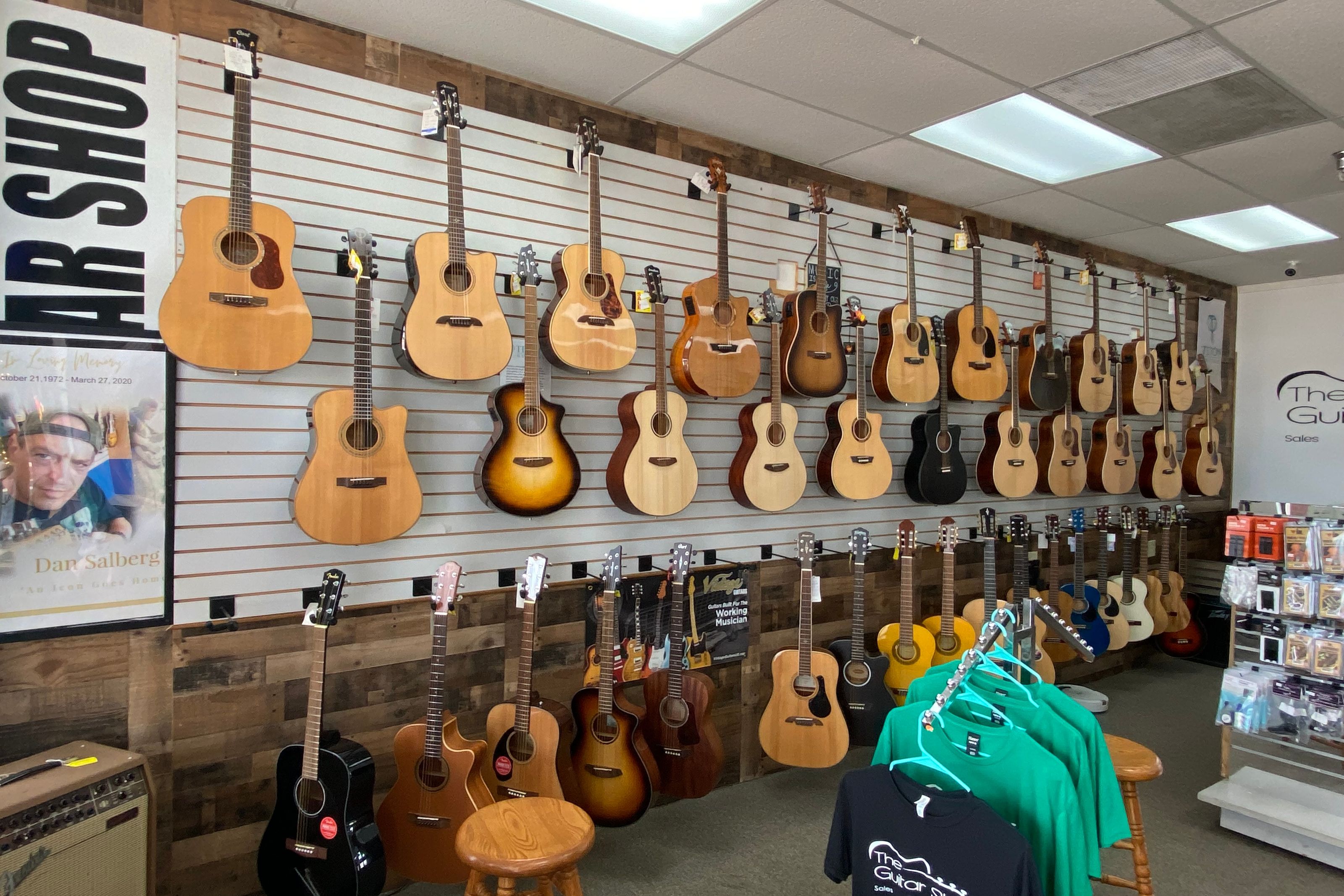 Wall of acoustic guitars over the reclaimed wood base, with shop t-shirts on the rack below