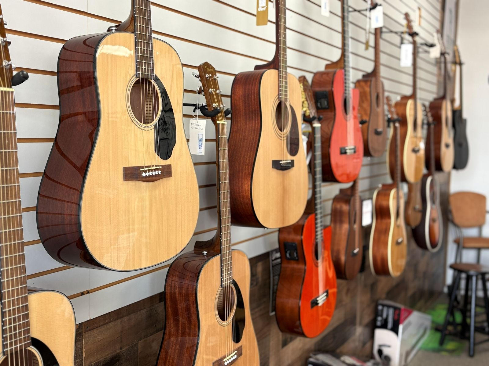 A natural-top acoustic dreadnought on the wall, guitars receding down the row behind it