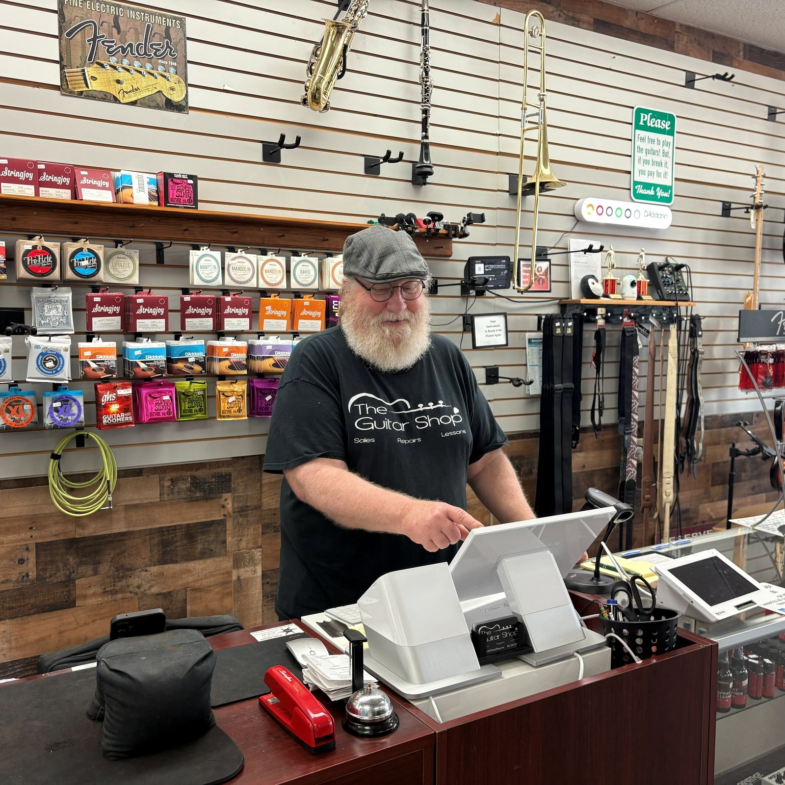 A staff member in a Guitar Shop t-shirt working behind the counter, string wall and accessories behind him