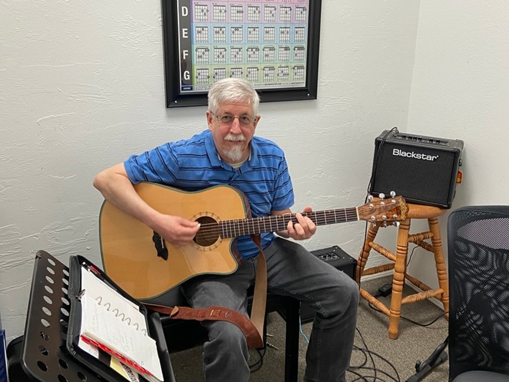 Tilden Barger playing an acoustic guitar in a lesson room with a chord chart on the wall