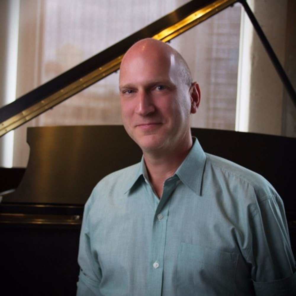Tim Oynes smiling in front of a grand piano by a bright window