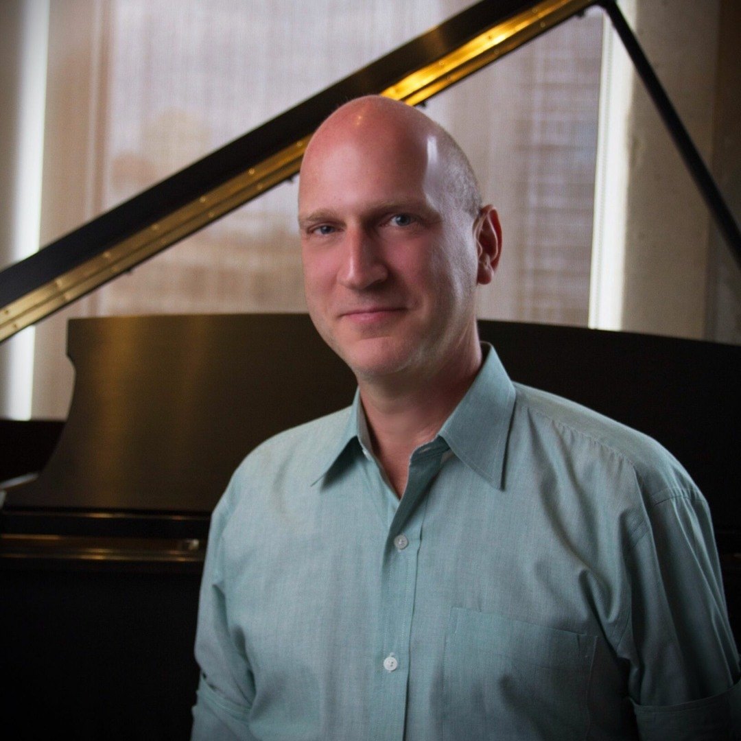Tim Oynes smiling in front of a grand piano by a bright window